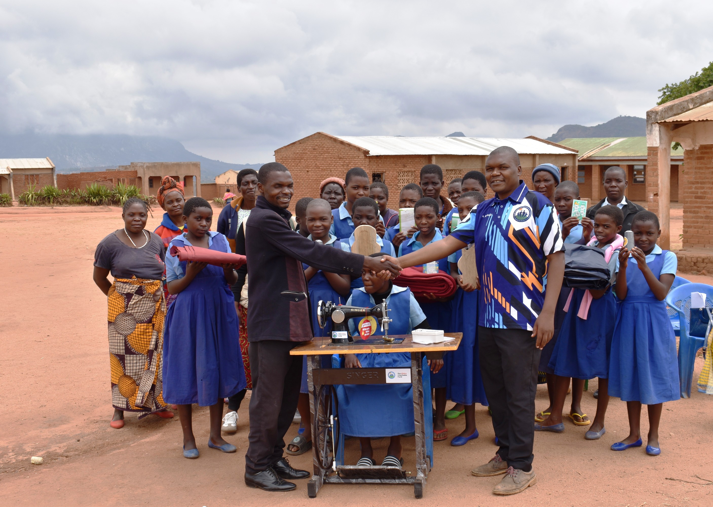 Bwanali (right) handing over the sewing machine and start-up materials to the Headteacher of Mwatonga Primary School as school girls and members of Mothers' grouplook on